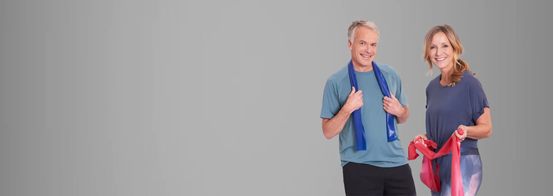 Ein Mann und eine Frau stehen vor einem grauen Hintergrund und halten jeder ein TheraBand in der Hand