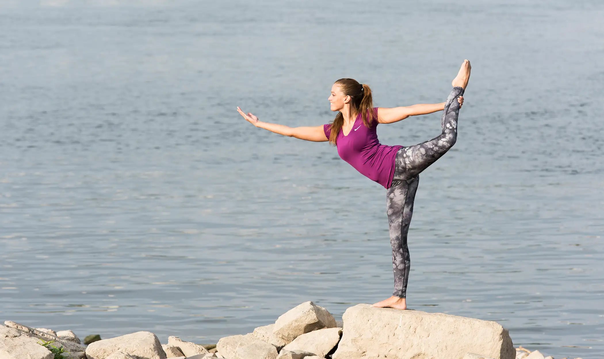 Frau balanciert auf einem Bein auf einem Felsen vor einem Gewässer.