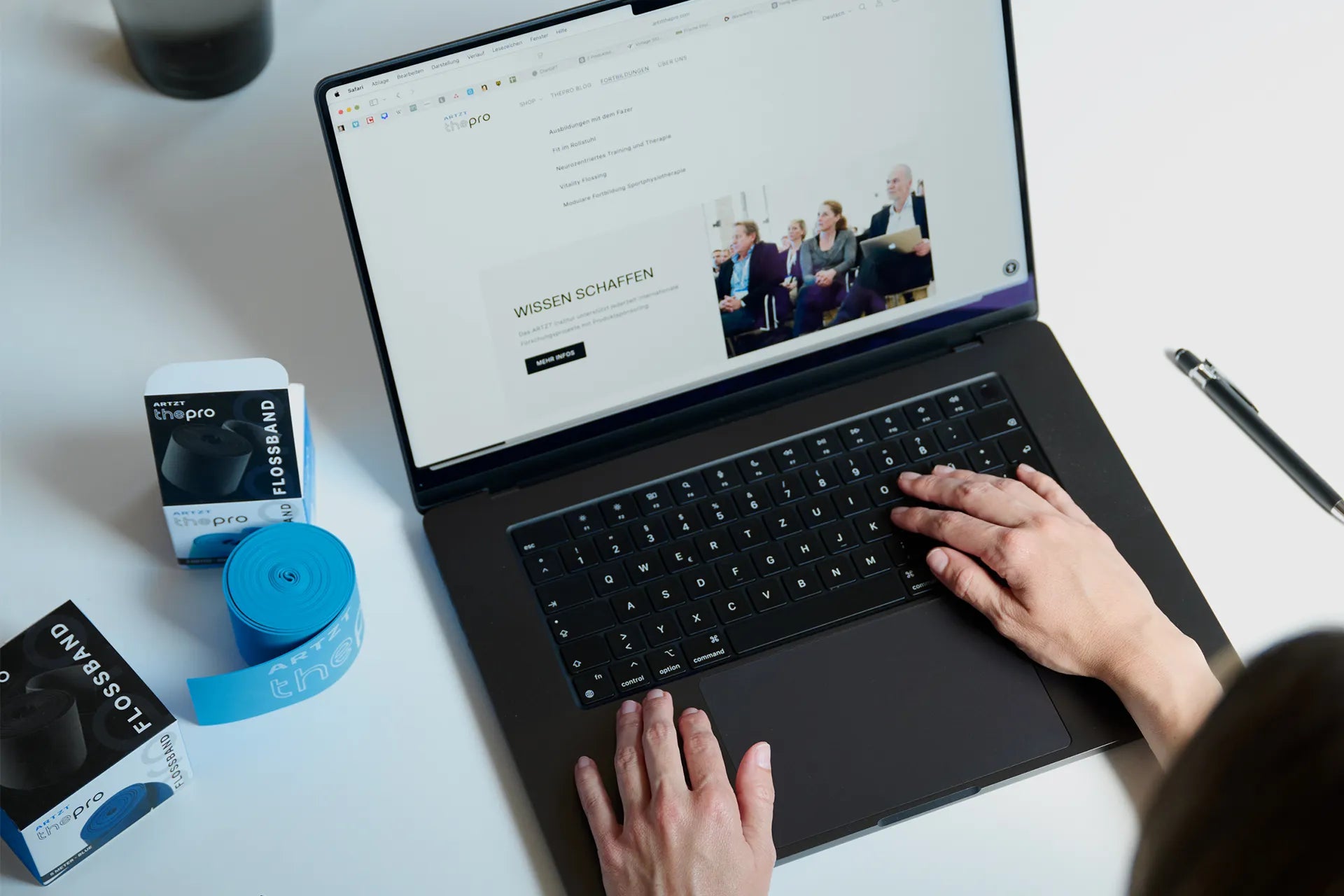 Zwei Hände ruhen auf einem geöffneten Laptop, der auf einem weißen Schreibtisch steht. Daneben liegt einARTZT thepro Flossband.