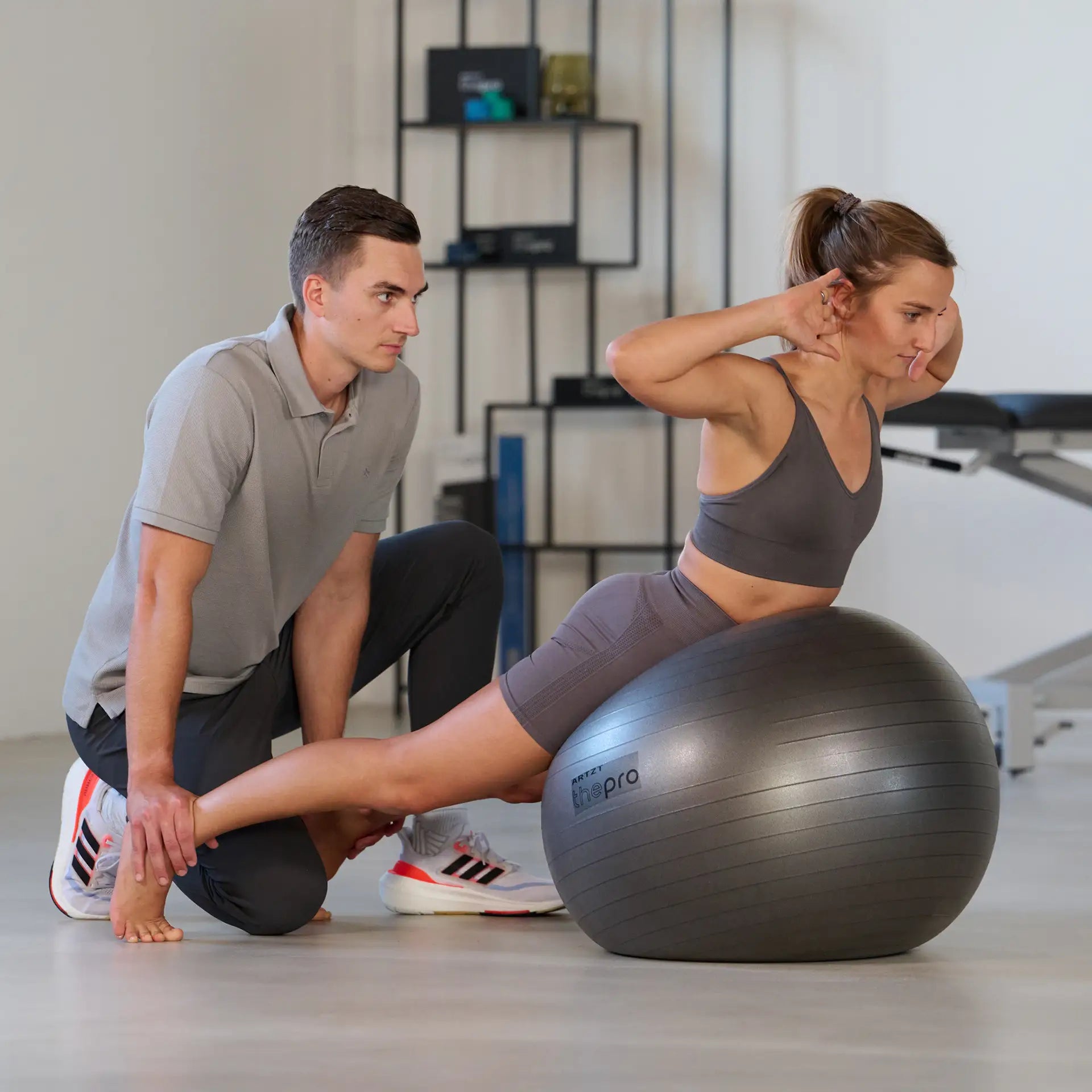 Sportlerin führt eine Rumpfstreck-Übung sitzend auf einem Gymnastikball der Marke ARTZT thepro aus, während ein Trainer sie seitlich begleitet und das Bein stabilisiert. Die Szene zeigt funktionelles Training zur Verbesserung von Rumpfstabilität, Haltung und Körperkontrolle.
