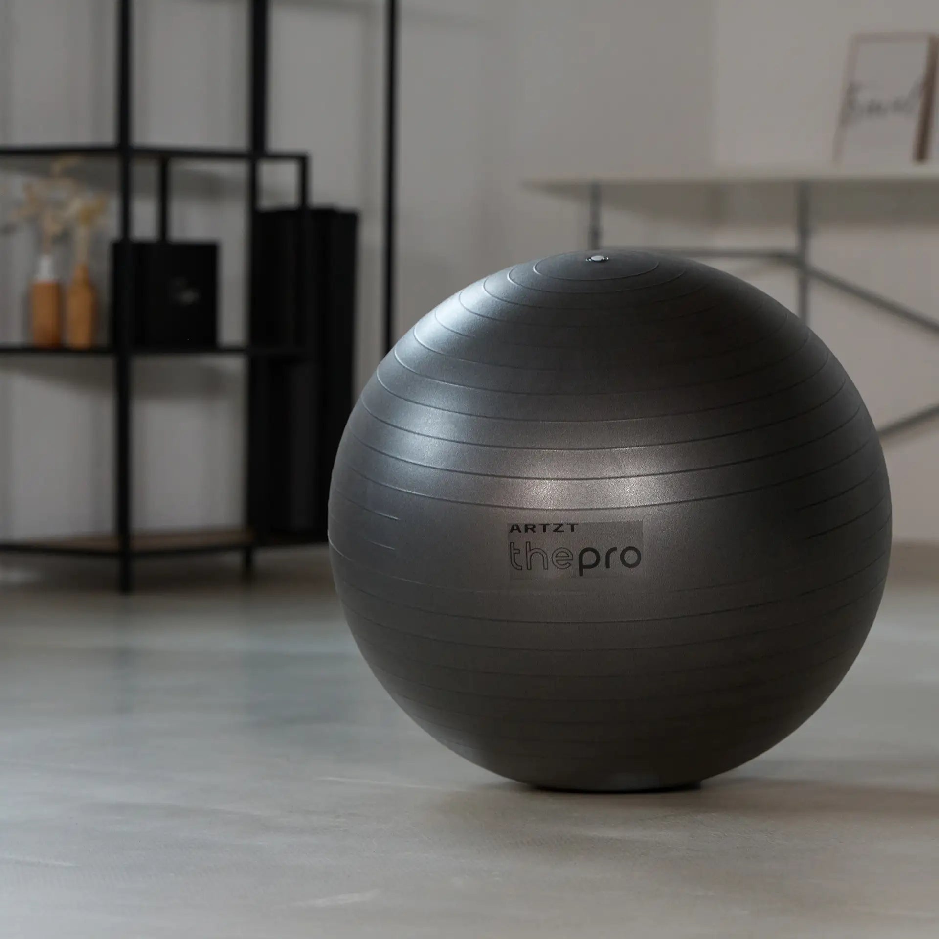 Grauer Gymnastikball der Marke ARTZT thepro steht frei auf dem Boden eines Trainingsraums; die gerippte Oberfläche und das aufgedruckte Logo sind gut sichtbar und zeigen den Ball als vielseitiges Trainings- und Therapiegerät.