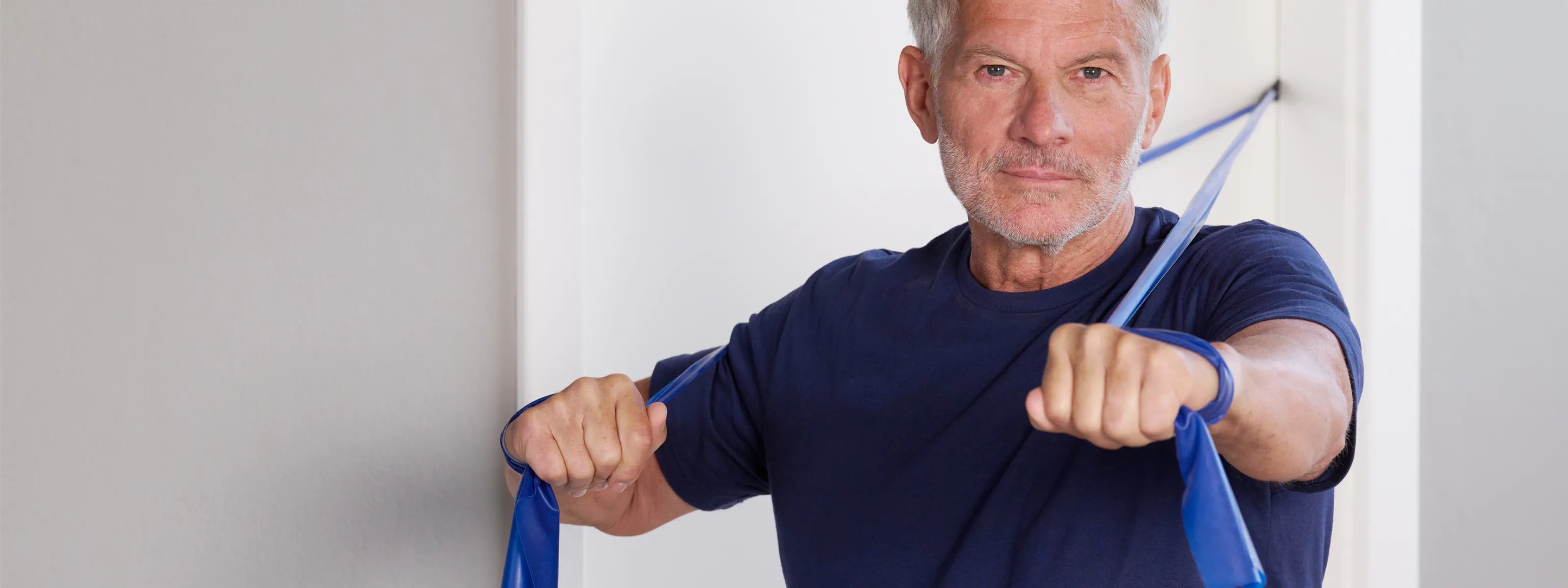 Mann macht eine TheraBand Übung, bei der er ein in die Tür eingeklemmtes Fitnessband mit beiden Armen nach vorne zieht.