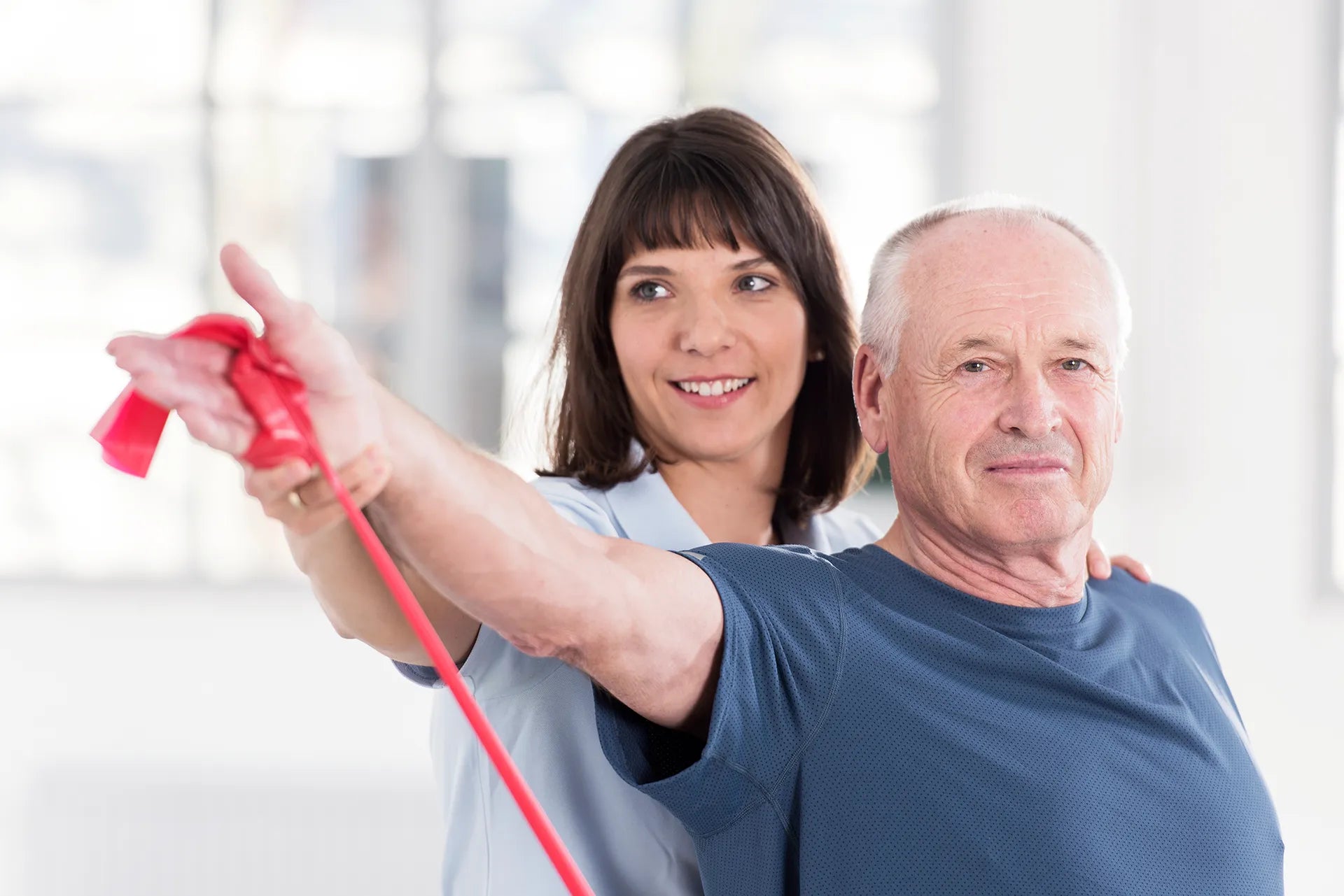 Älterer Mann führt unter Anleitung einer Therapeutin eine Schulterübung mit rotem Theraband im Innenraum durch, Arm seitlich ausgestreckt zur Kräftigung und Mobilisation.