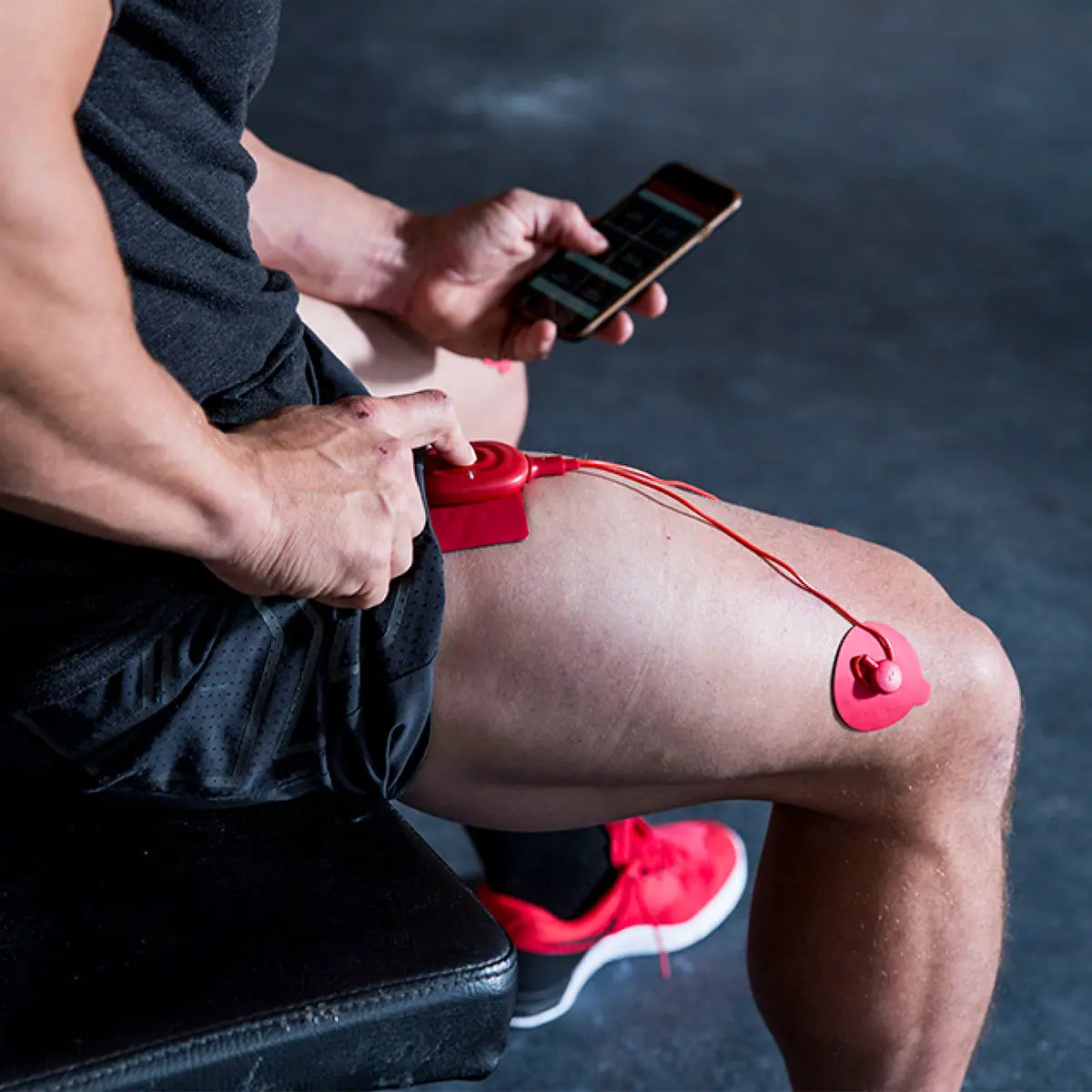 Ein Mann trägt Therabody PowerDot UNO Muskelstimulatoren auf dem rechten Oberschenkel und hält ein Smartphone in der linken Hand.