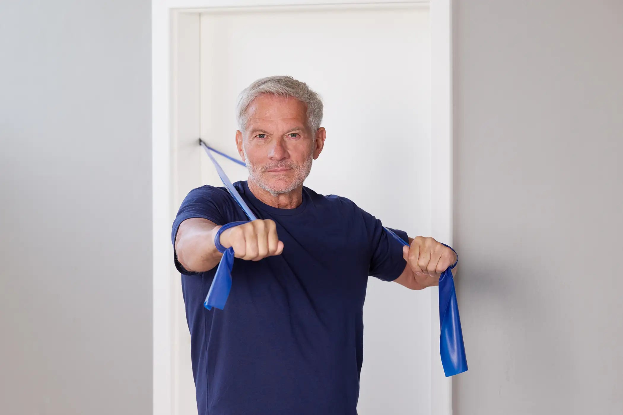 Ein Mann steht vor einer weißen Tür und trainiert seine Brust mit einem blauen TheraBand Fitnessband für mehr Longevity