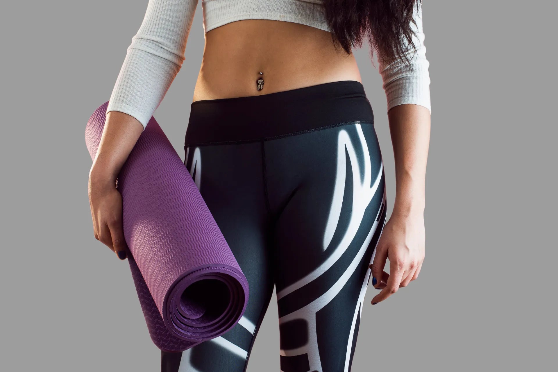 Frau im Sportoutfit hält eine zusammengerollte Yogamatte in der Hand; Nahaufnahme von Bauch- und Beinbereich mit schwarzer Trainingsleggings vor neutralem Hintergrund, Symbolbild für Fitnesstraining und Bewegung.