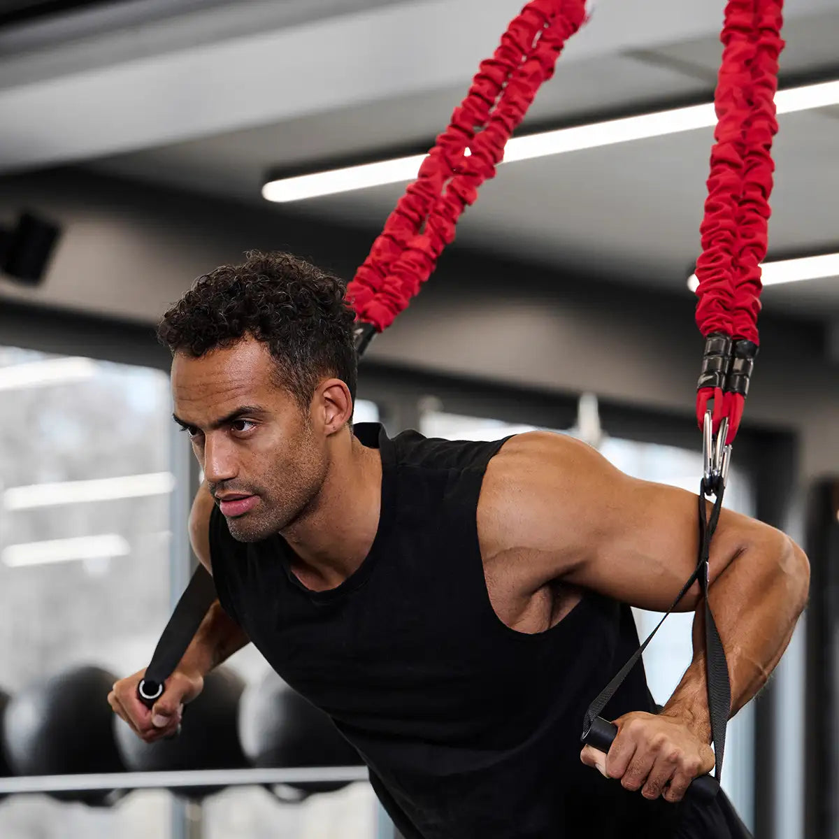 Sportler führt eine Übung mit dem 4D PRO Bungee Schlingentrainer aus, gehalten an zwei roten elastischen Lambdabändern mit Griffen in einem Fitnessstudio.