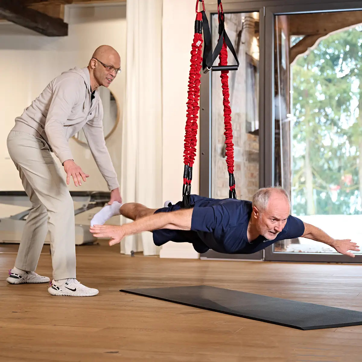 Therapeut begleitet eine Übung mit dem 4D PRO Bungee Schlingentrainer, während ein Mann mit dem Körper horizontal in den elastischen Seilen trainiert.