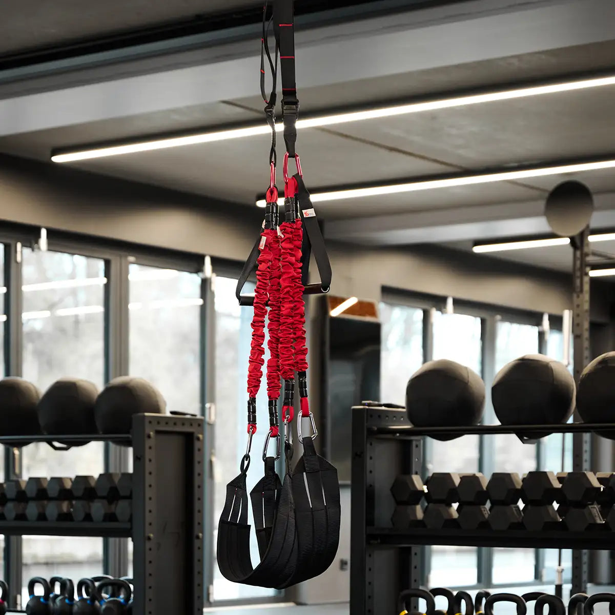 4D PRO Bungee Schlingentrainer mit roten Bungee-Seilen und schwarzen Schlaufen, aufgehängt an einer Deckenbefestigung in einem Fitnessstudio.