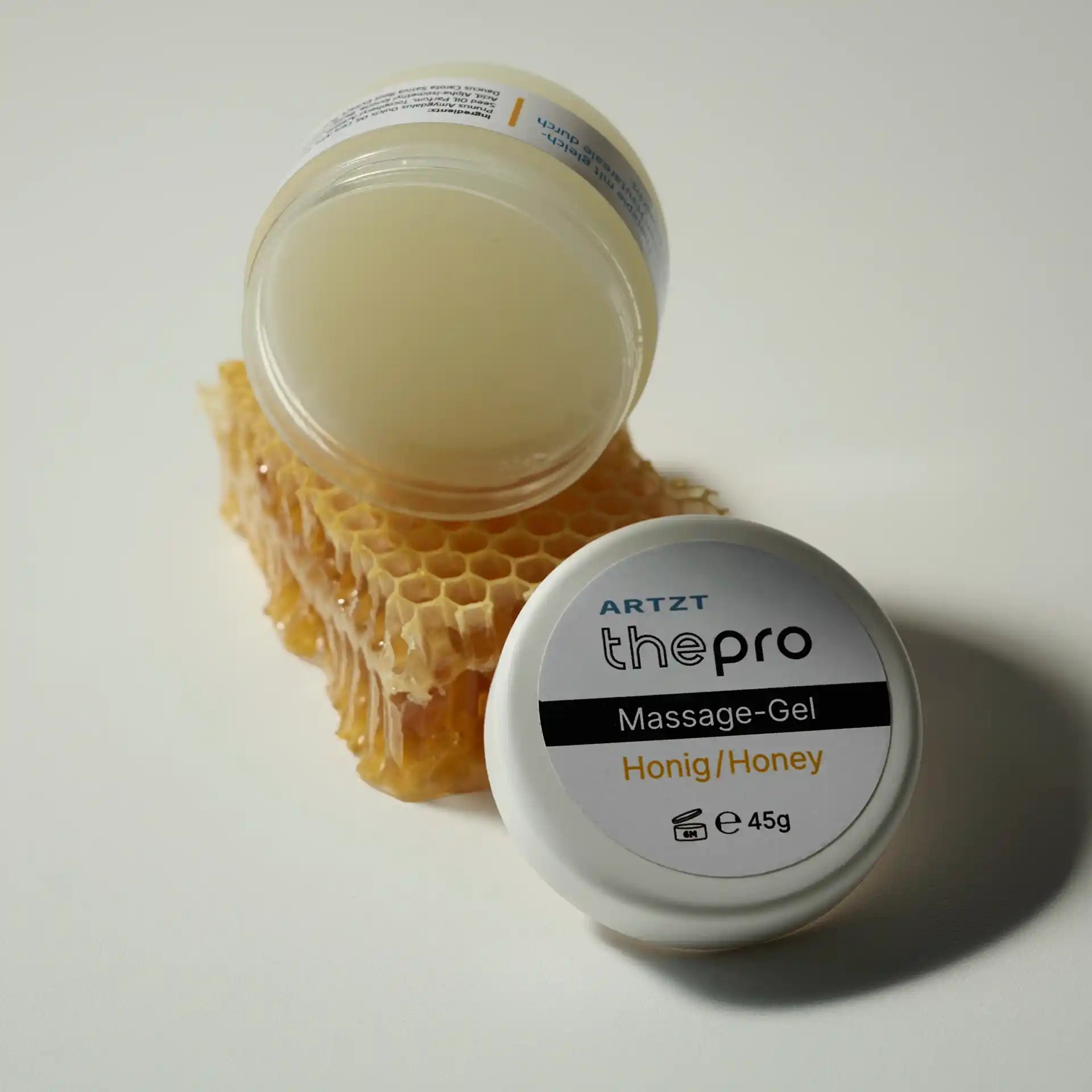 Eine geöffnete Dose ARTZT thepro Massage-Gel Honig steht hochkant auf einem Stück Bienenwachs. Daneben lehnt der Deckel mit der Beschriftung nach oben.