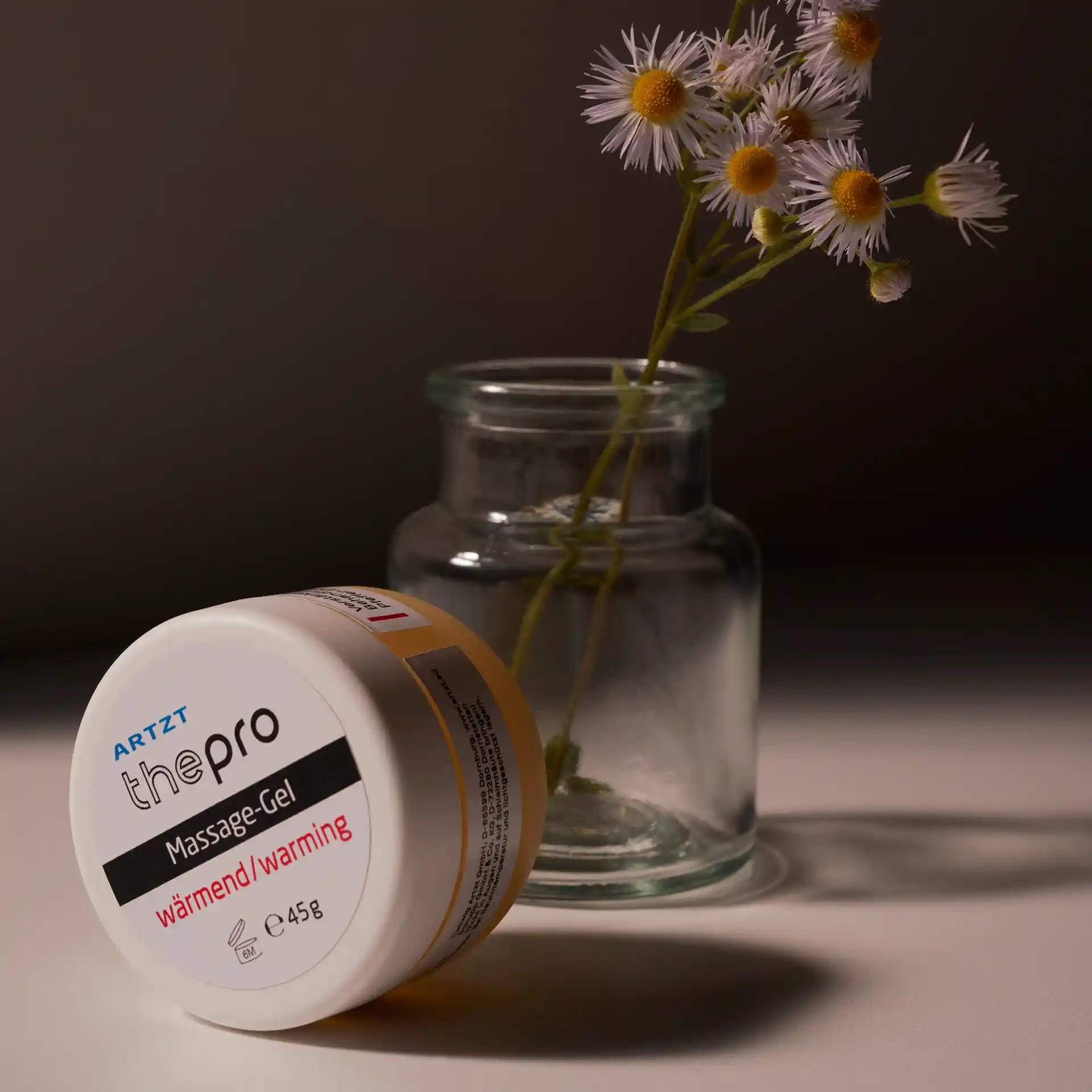 Eine Dose ARTZT thepro Massage-Gel wärmend steht hochkant auf einer hellen Oberfläche. Dahinter steht ein Glas mit einem kleinen Strauß weißer Blumen mit gelber Blüte.