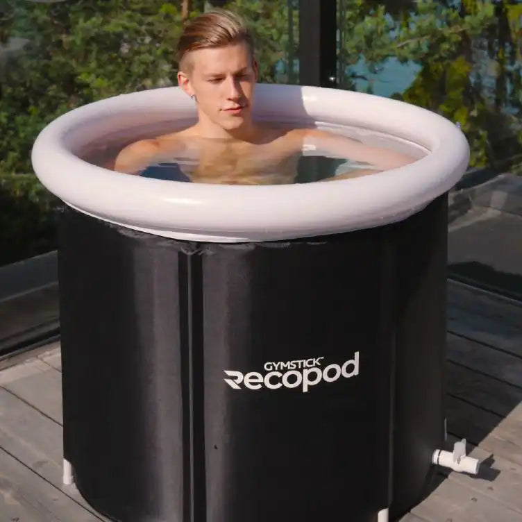 Person sitzt bis zur Brust in der RecoPod Eistonne von Gymstick im Außenbereich auf einer Holzterrasse, die schwarze Tonne ist mit weißem Rand gefüllt mit Wasser, Ablassventil seitlich sichtbar.