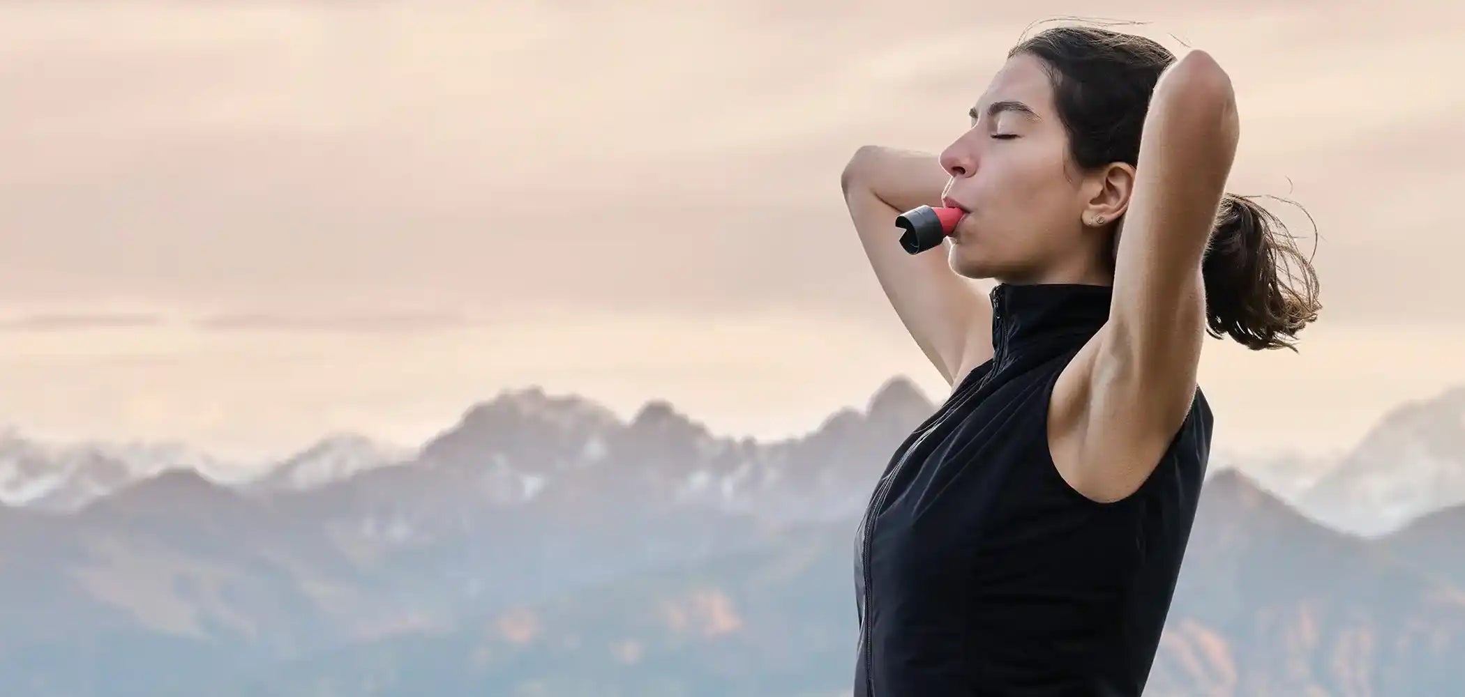 Eine Frau steht vor einen Bergpanorama, verschränkt die Arme hinter dem Kopf, hat die Augen geschlossen und trägt im Mund einen ARTZT neuro Atemtrainer.