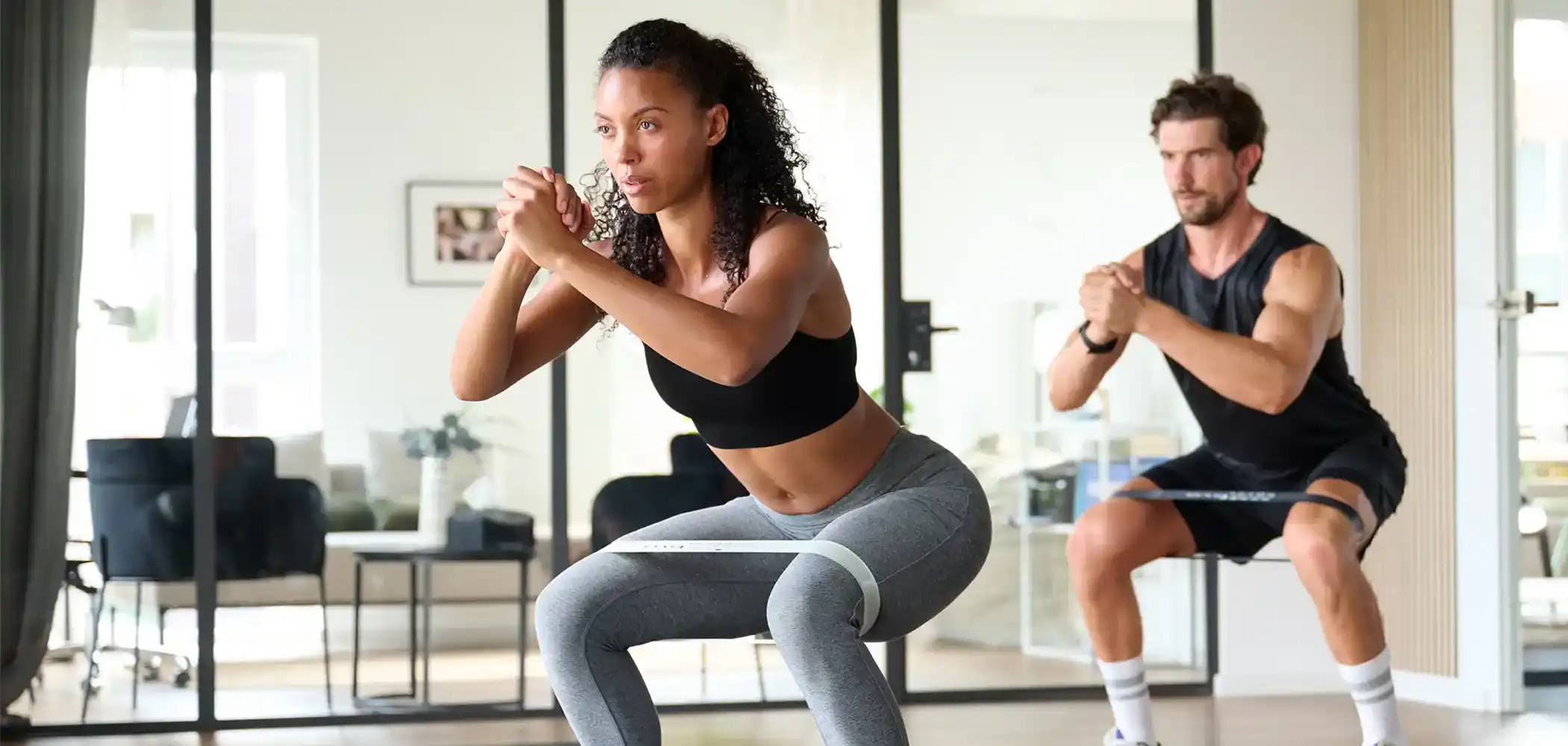 Eine Frau und ein Mann machen in einem Wohnzimmer Squats mit jeweils einem ARTZT thepro Loop Band um die Oberschenkel gelegt.