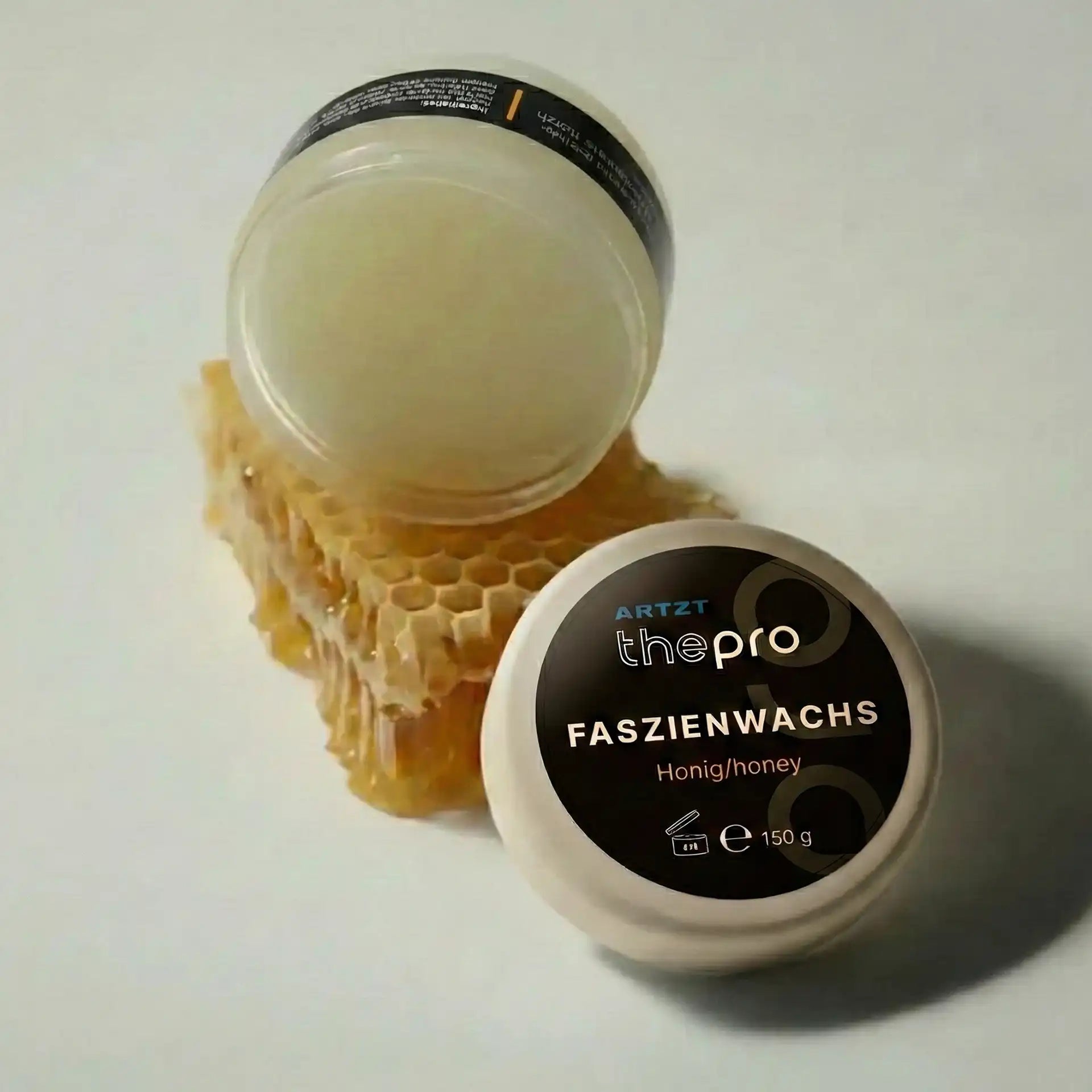 Geöffnete Dose Faszienwachs Massagegel „Honig/Honey“ von ARTZT thepro mit 150 ml Inhalt, weißer Deckel daneben liegend, Dose auf einem Stück Bienenwabe platziert, honigfarbenes Gel sichtbar.