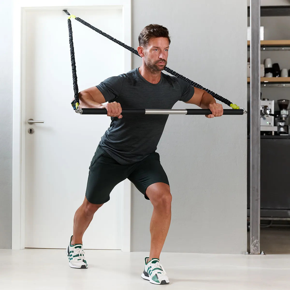 Mann führt eine Kraftübung im Ausfallschritt mit einem HRT Fitnessband und Trainingsstange von ARTZT thepro in einem Innenraum aus.