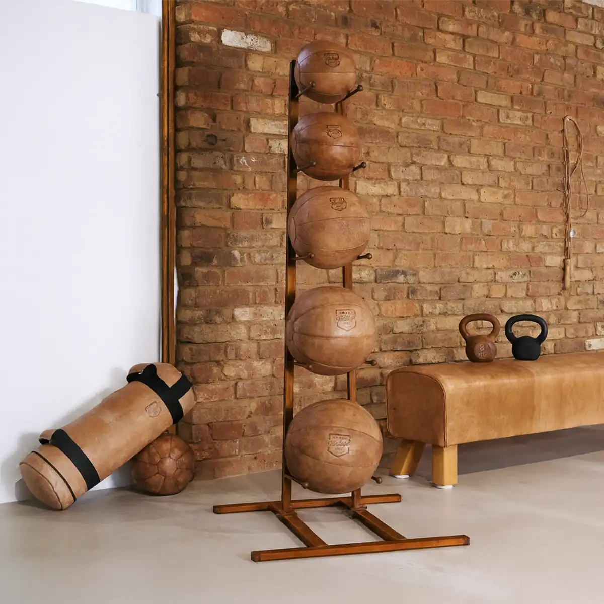 Ein ARTZT Vintage Medizinball Ständer bestückt mit fünf Lederbällen steht vor einer Ziegelwand.