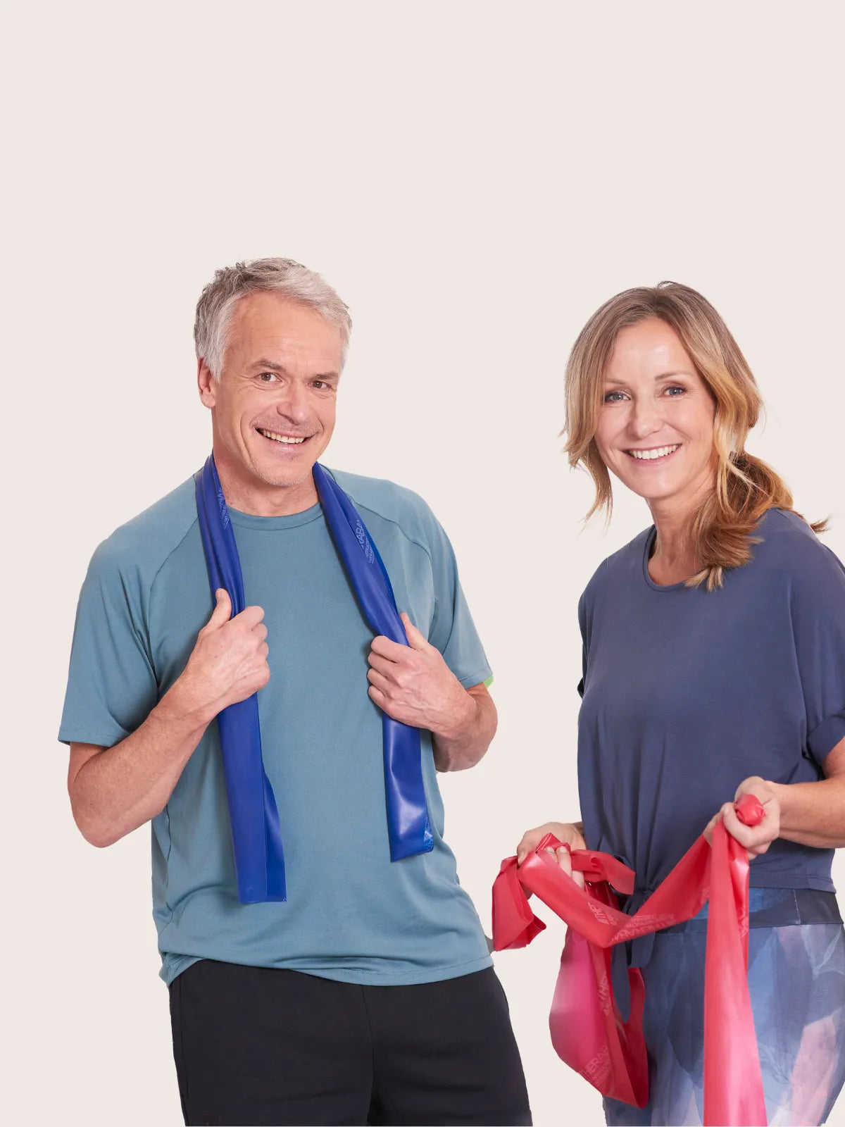 Ein Mann und eine Frau stehen vor einem beigen Hintergrund, jeweil ein TheraBand in den Händen.