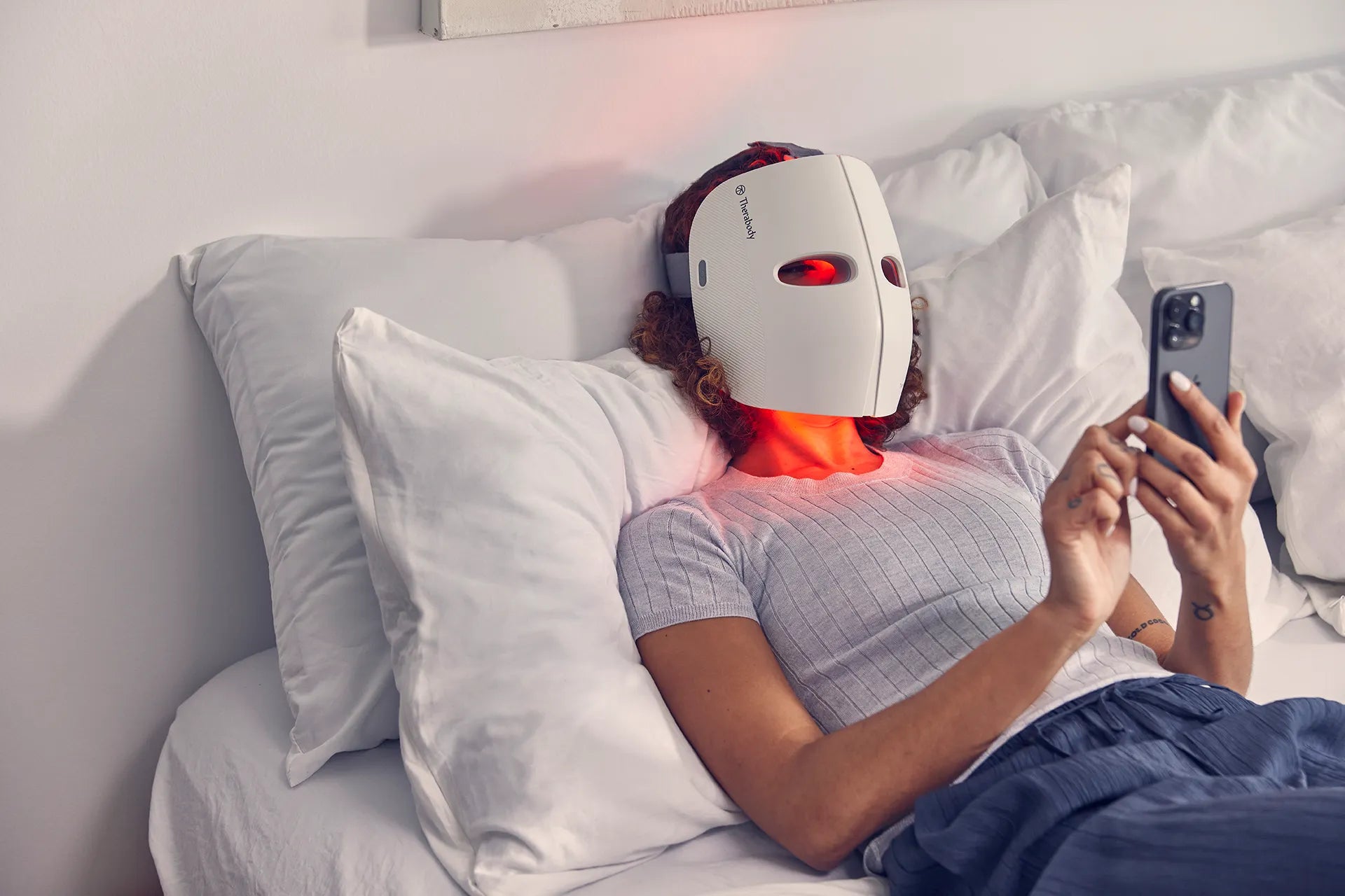Frau liegt im Bett mit einem Smartphone in der Hand und trägt auf dem Gesicht eine Therabody TheraFace Mask LED-Maske.