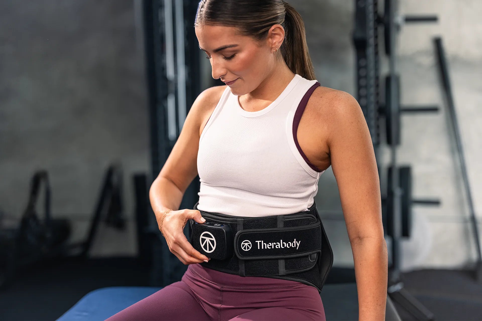 Frau sitzt in einem Trainingsraum und trägt einen Therabody ThermBack LED um die Hüfte.