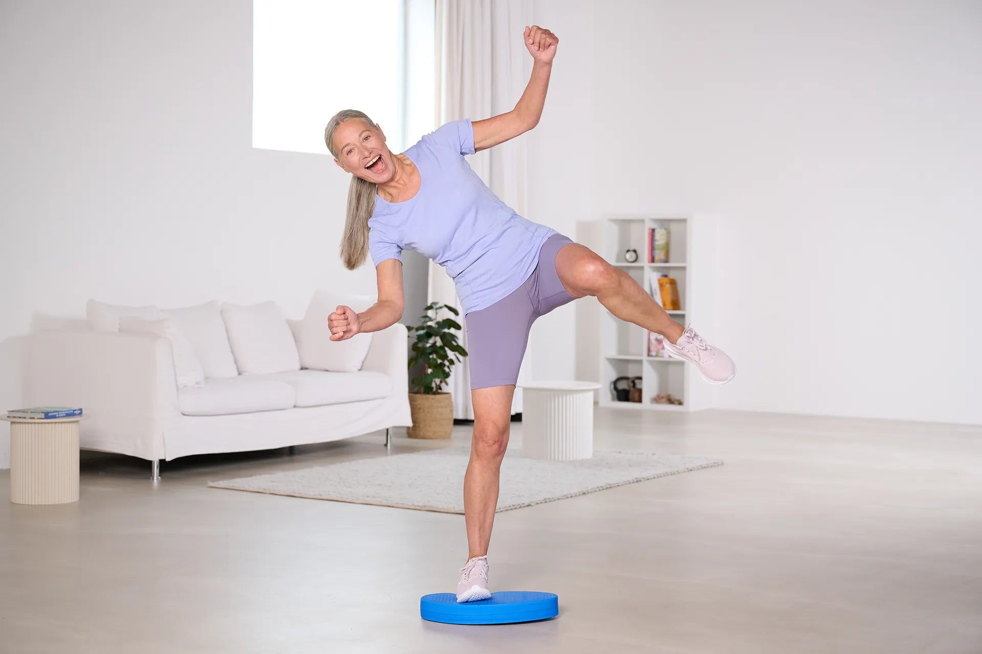 Eine Frau steht lachend mit dem rechten Fuß auf einem blauen Balance Pad der Marke TheraBand, während sie das linke Bein zur Seite spreizt, als ob sie kurz davor sei, das Gleichgewicht zu verlieren.