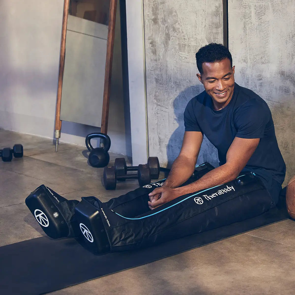 Ein Mann trägt Therabody JetBoots Kompressionsstiefel an seinen Beinen und lehnt dabei im Sitzen lächelnd an einer Wand.