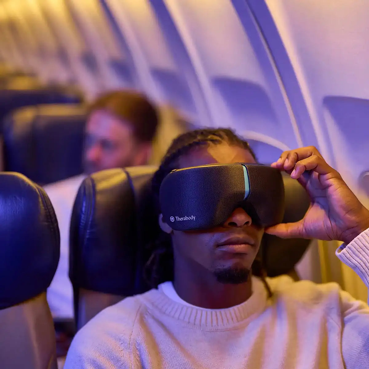 Mann sitzt im Flugzeug und bedient mit der linken Hand ein Therabody SmartGoggles Wearable.
