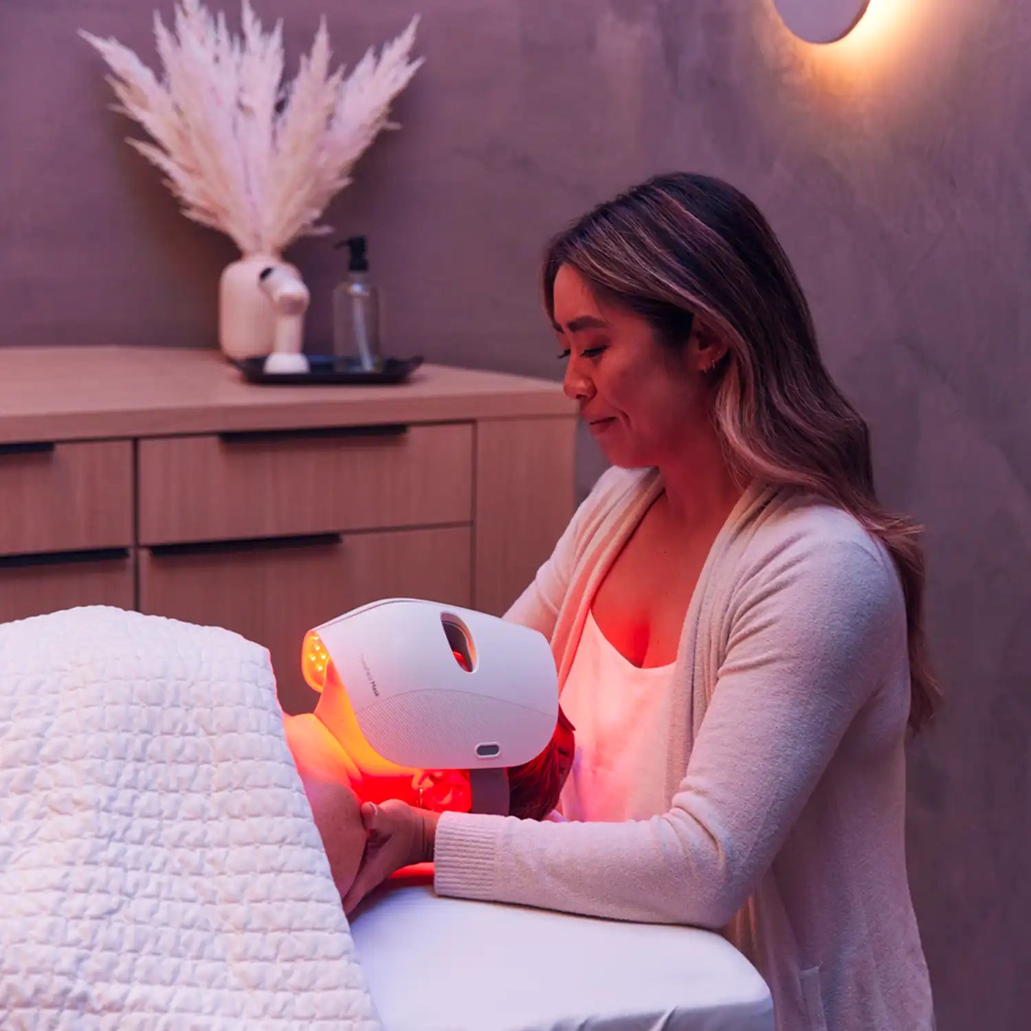 Frau steht hinter einem Mann, der auf einer Massageliege liegt und eine Therabody TheraFace Mask LED-Maske trägt.